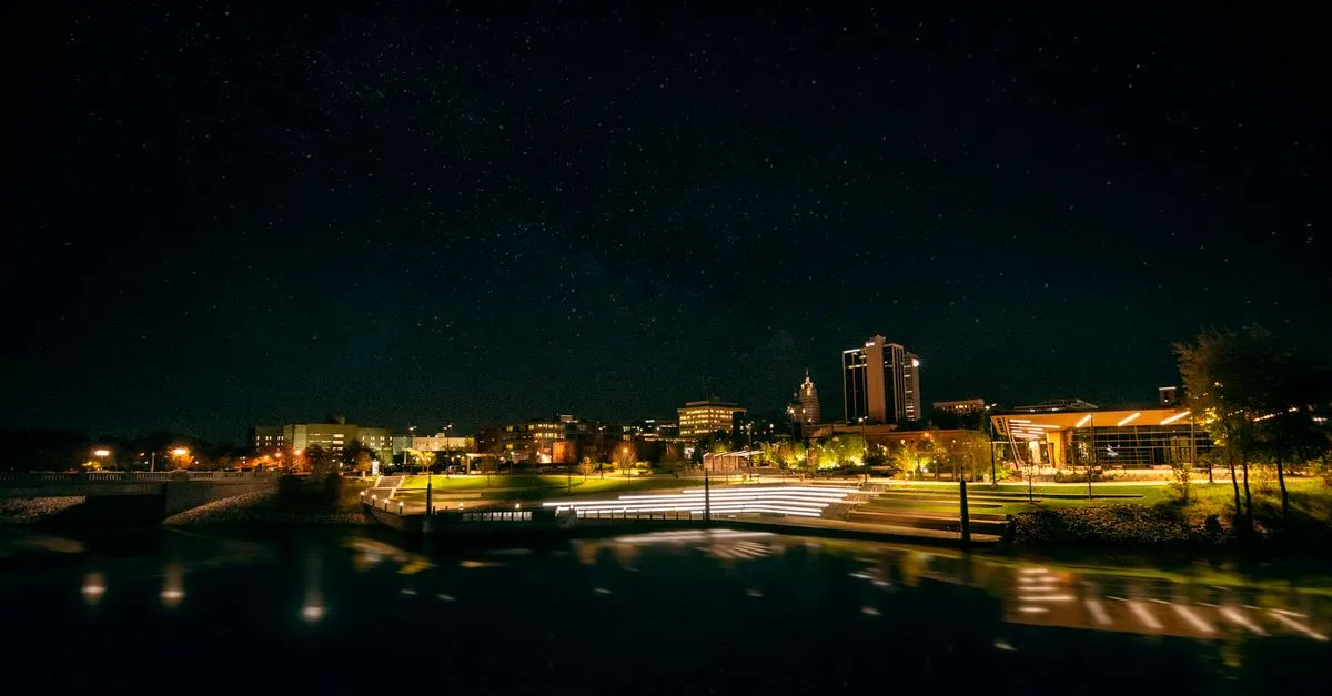 Night view of the Fort Wayne, Indiana skyline lit up beyond a riverfront park under a clear, starry sky.