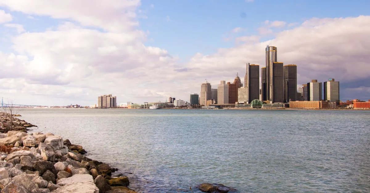 The downtown Detroit skyline seen across the Detroit River on a clear day, with the Renaissance Center towers above the waterfront and a rocky shoreline in the foreground.