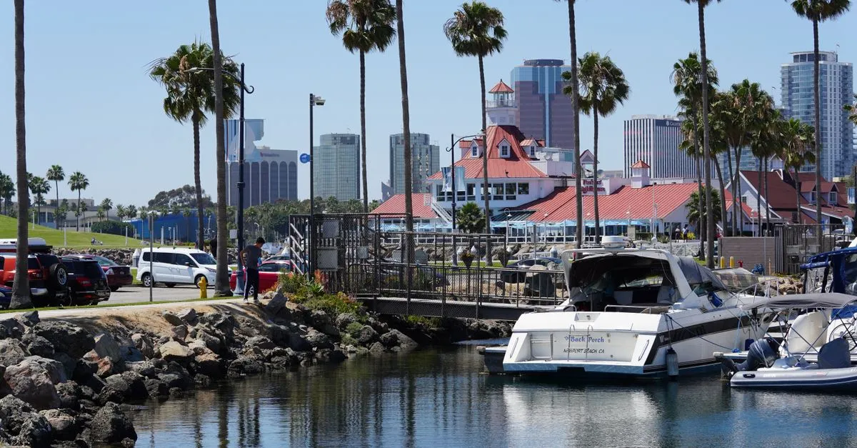 The Long Beach, California skyline rising behind Rainbow Harbor, framed by palm trees and a red-roofed waterfront restaurant on a clear day.