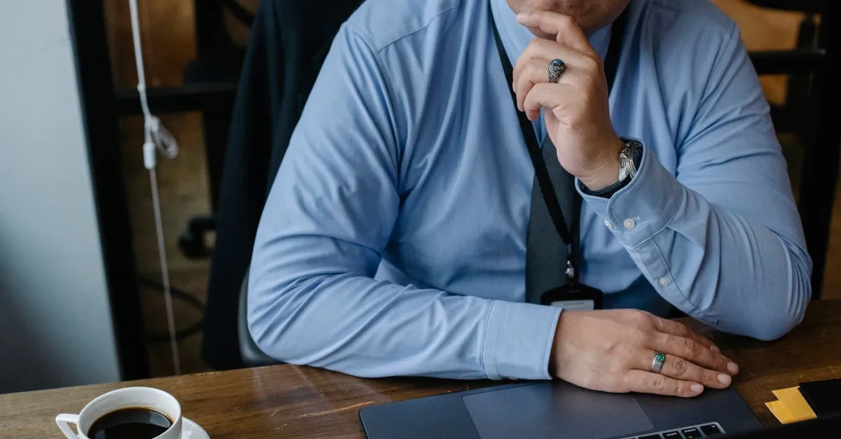 A business owner sits at a desk with a coffee and a laptop, hand on chin, thinking about what people find when they look the company up.