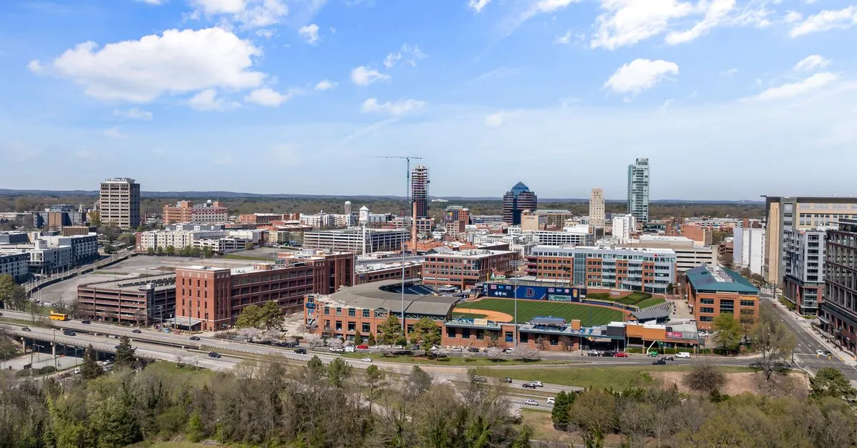 Aerial view of downtown Durham, North Carolina, with the skyline rising behind Durham Bulls Athletic Park and the American Tobacco district on a sunny day.