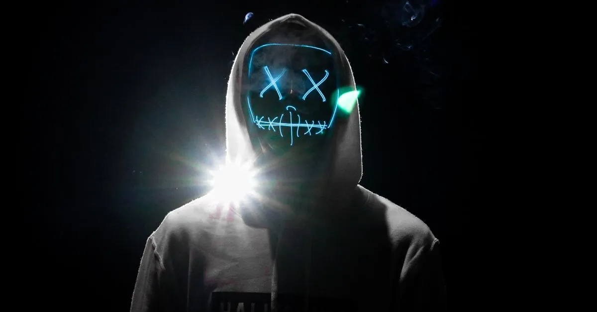 A hooded figure in a glowing LED mask standing in the dark, standing in for the negative SEO attacker most owners imagine.