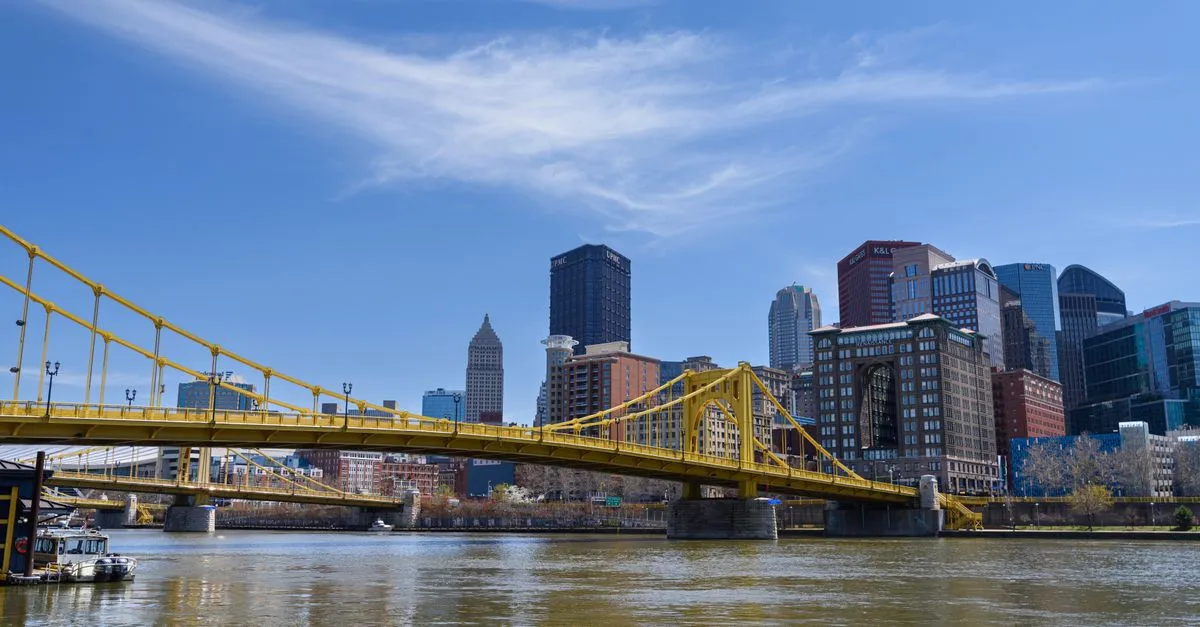 A yellow steel bridge over the Allegheny River with the downtown Pittsburgh skyline and UPMC tower behind it on a clear day.