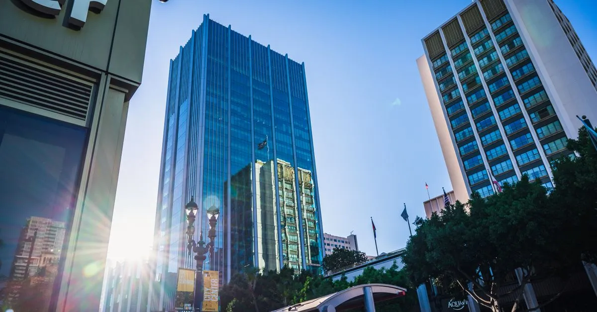 A modern glass office tower rising above a downtown business district under a clear blue sky.