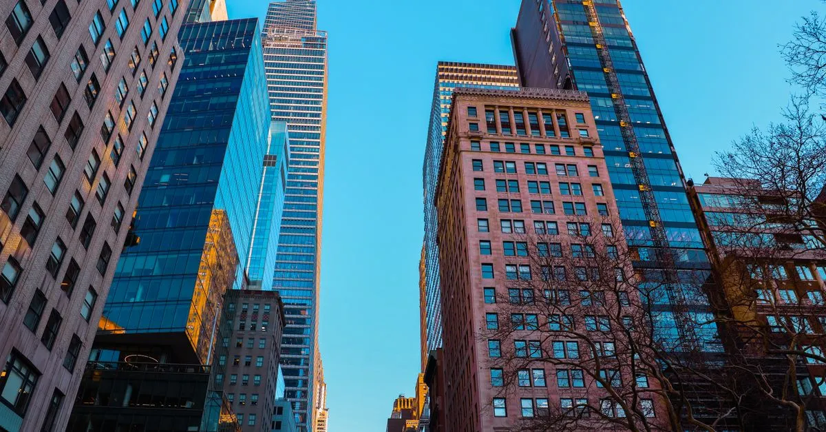 A low-angle view up a downtown financial district, with glass and stone office towers rising against a clear blue sky.