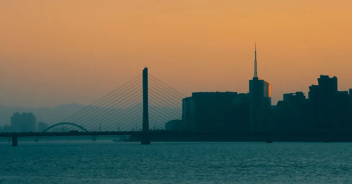 A cable-stayed bridge and downtown skyline silhouetted against a hazy orange sky at dusk over the water.