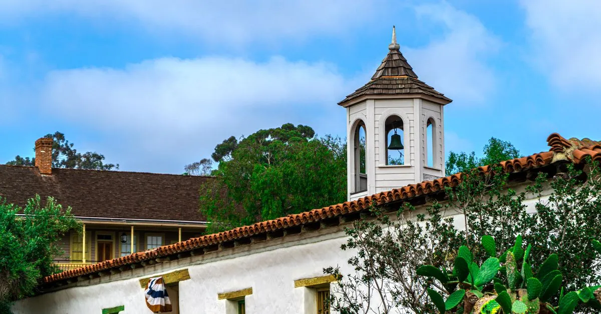 A historic California mission bell tower with a red tile roof and white adobe walls against a bright blue sky.