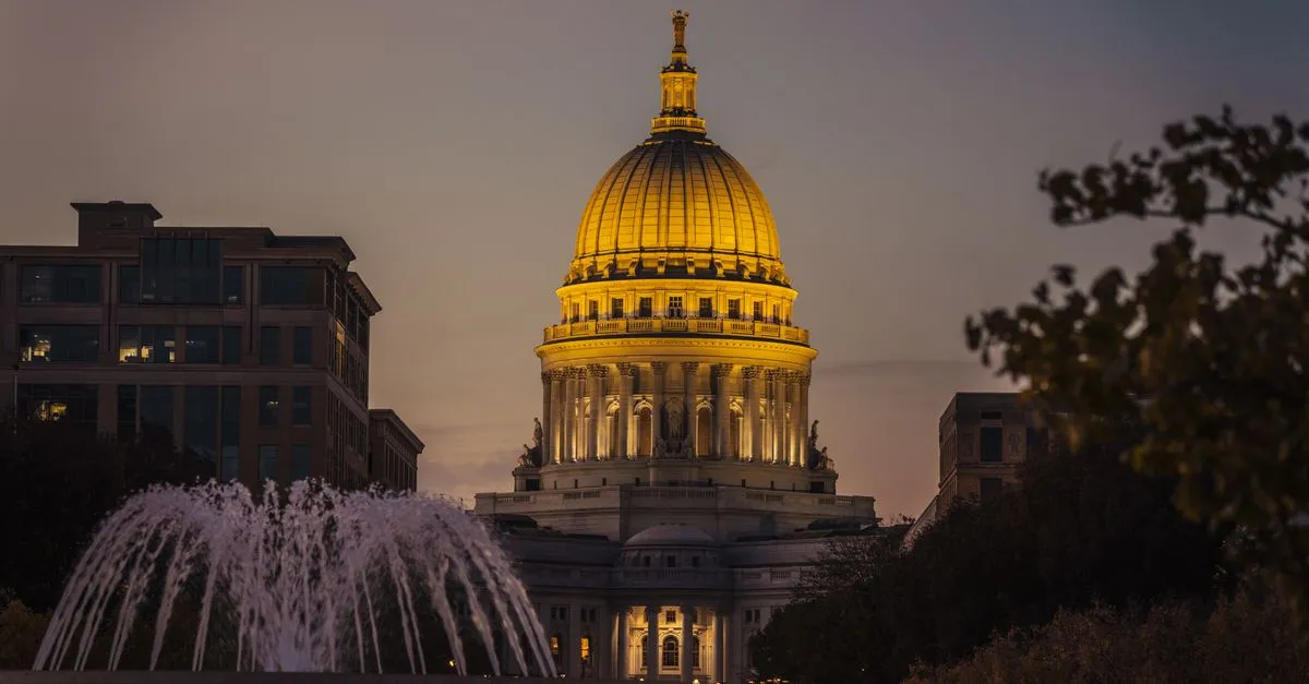 The golden dome of the Wisconsin State Capitol lit at dusk in downtown Madison, with a fountain spraying in the foreground.