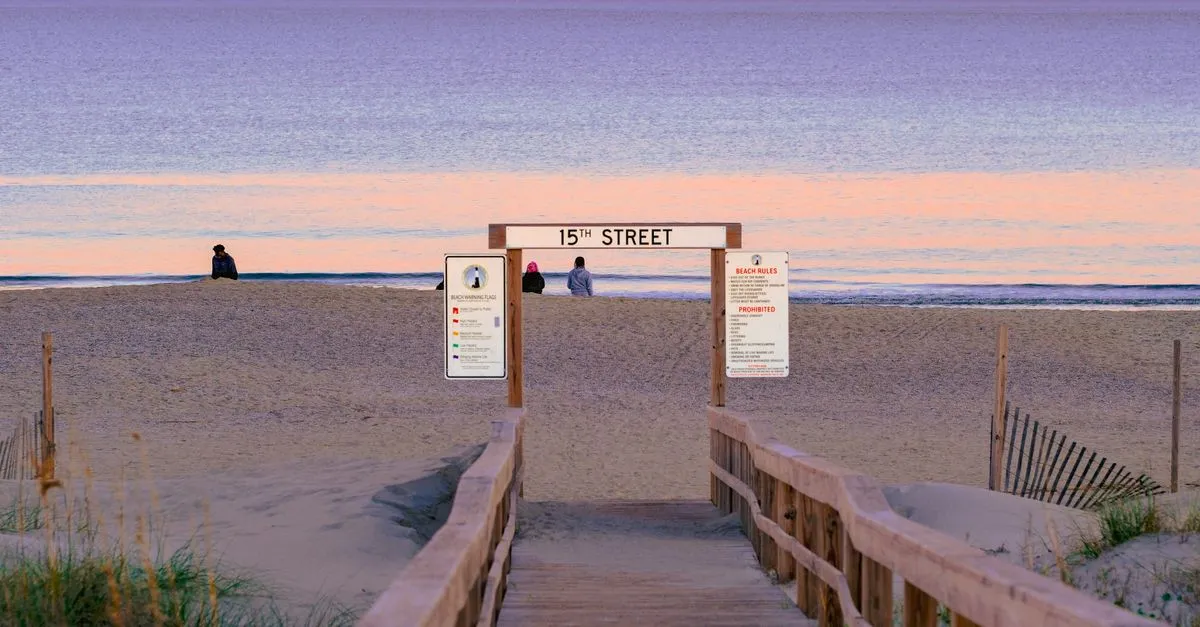 A wooden boardwalk ramp leading up to a wide sandy beach at a 15th Street access point, with beach warning flag and rules signs at sunset.