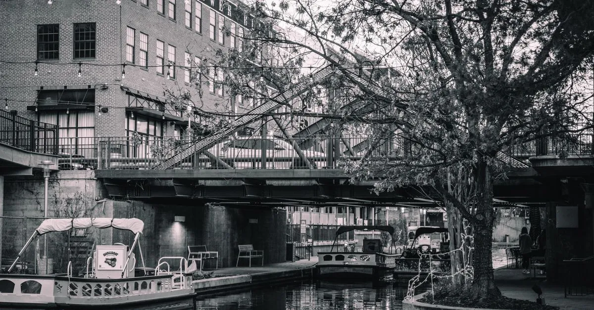 Flat-bottom tour barges and string lights along the San Antonio River Walk canal beneath a stone bridge, the tourist heart of the local market.