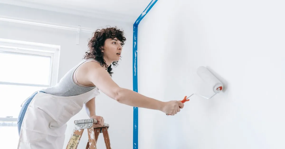 A painter on a step ladder rolls white paint onto a taped interior wall, the kind of pro painting company SEO helps get found.