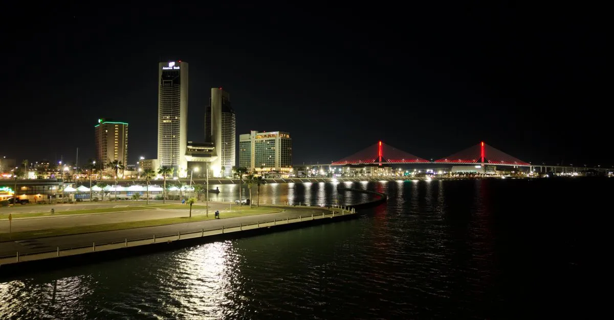 The Corpus Christi skyline lit up at night beside the illuminated Harbor Bridge over the bay.