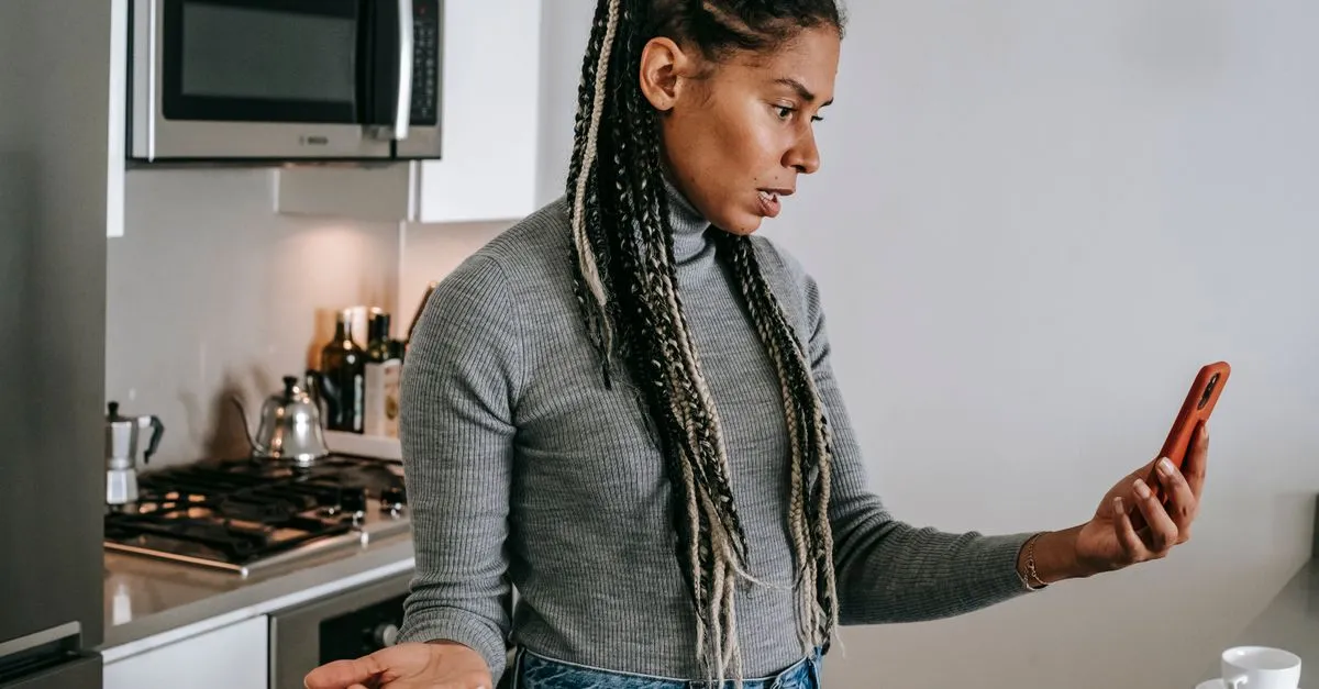 A woman standing in a kitchen looking confused and surprised at the phone in her hand.