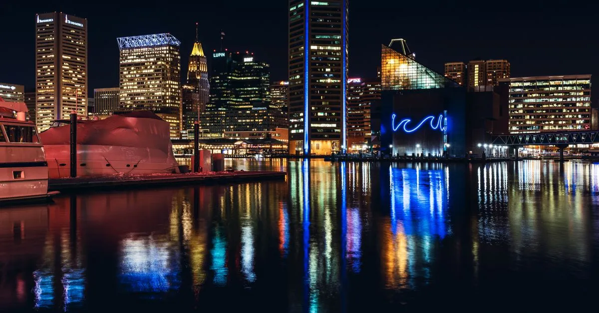 The Baltimore skyline lit up at night, its downtown towers reflecting across the Inner Harbor water, the dense local market where the map pack is won.