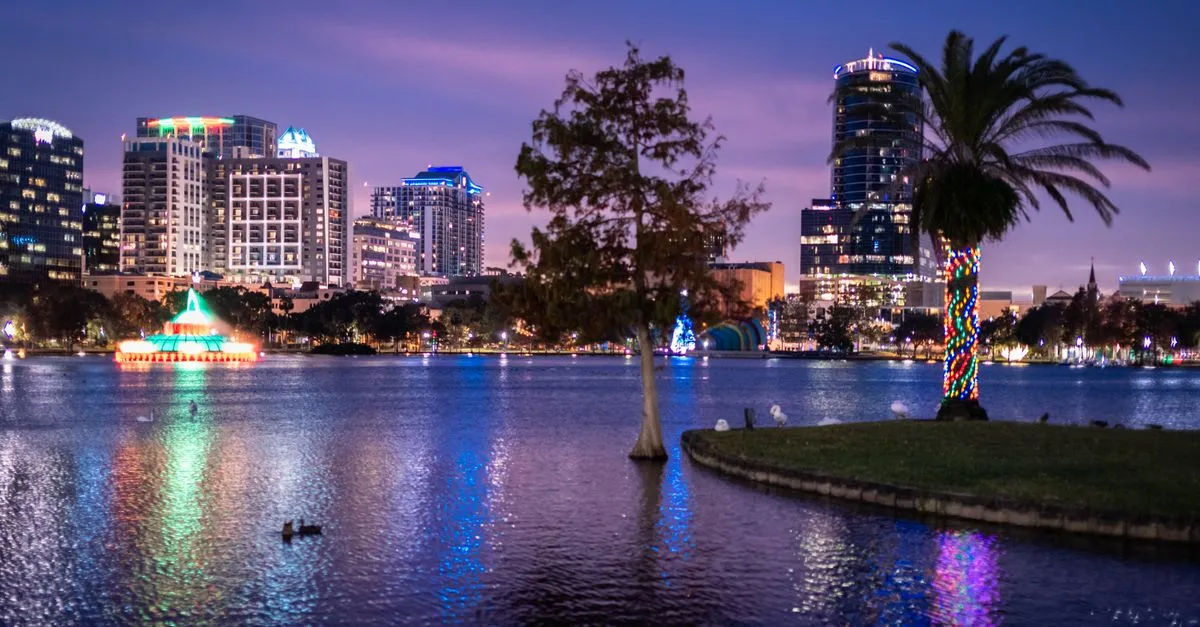 The downtown Orlando skyline at twilight reflected in Lake Eola, with a lit palm tree and holiday lights along the waterfront.