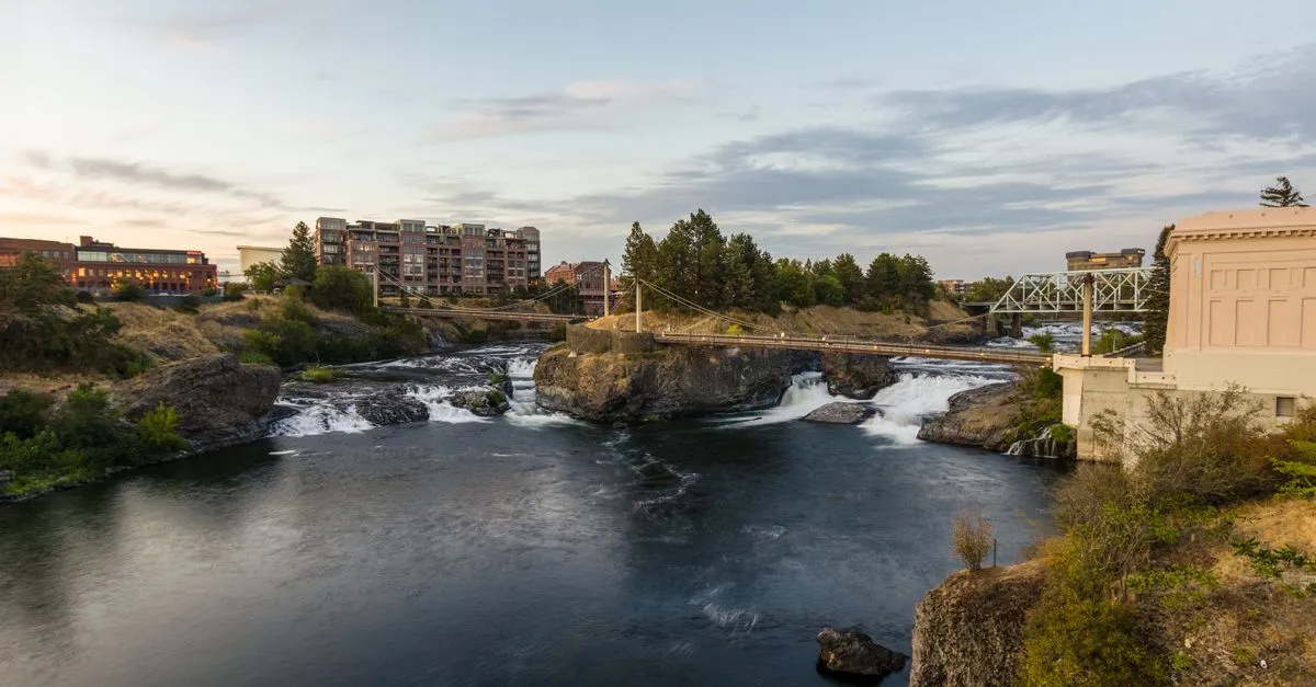 A view of Spokane Falls on the Spokane River at sunset, with a footbridge and downtown Spokane buildings in the background.