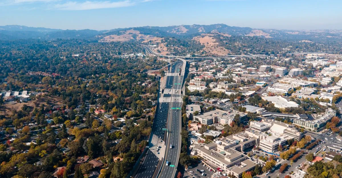 Aerial view of a Bay Area suburb where tree-lined homes meet a commercial strip and a freeway, with dry golden hills behind, the kind of dense local market the map pack is built for.
