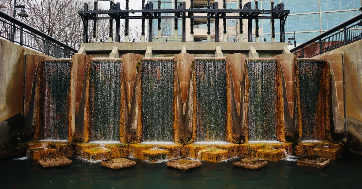 Water cascading over a stepped concrete weir along a downtown canal, with glass office buildings rising behind it.