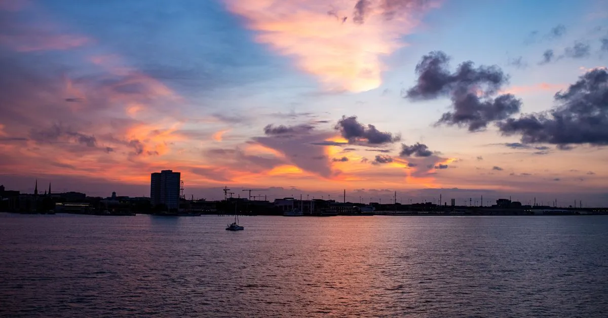 A waterfront city skyline silhouetted against a vivid sunset, with a lone sailboat on the calm water in the foreground.