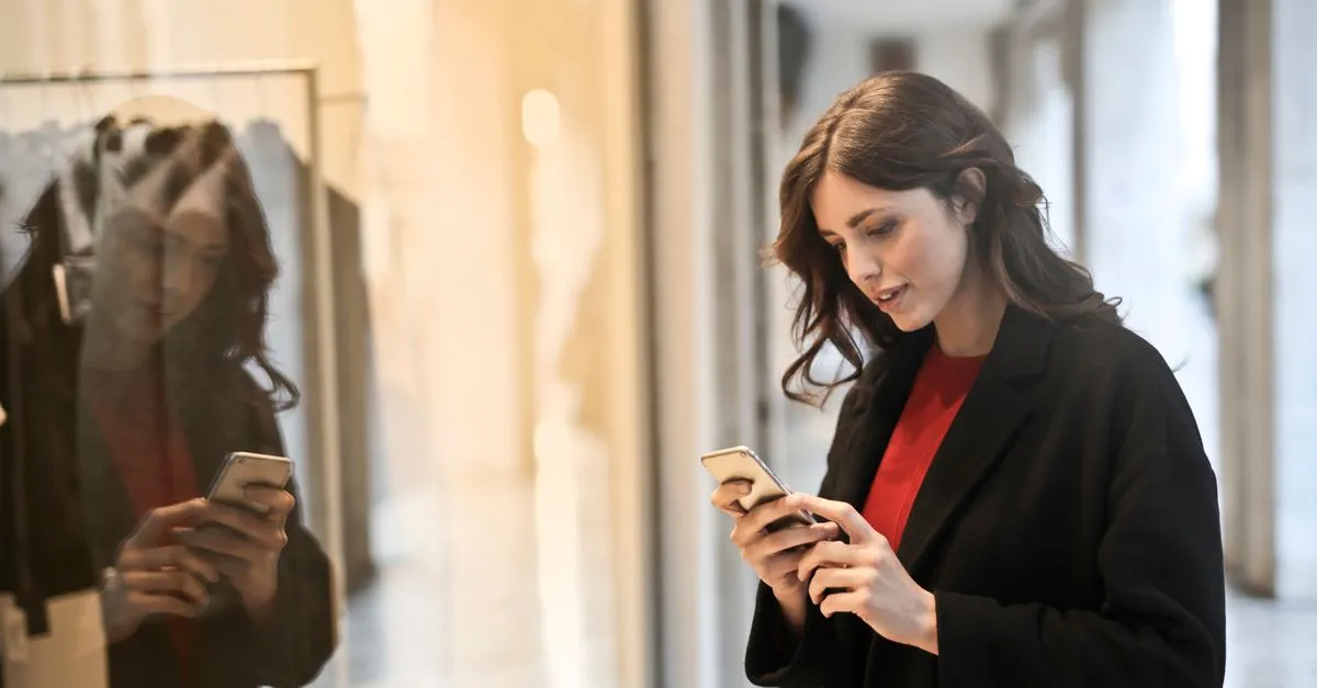 A woman in a dark coat checking her smartphone beside a shop window, her reflection visible in the glass.
