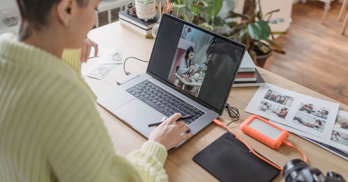 A person editing photos on a laptop with a stylus, next to printed photo sheets, an external drive, and a camera, showing image editing work.