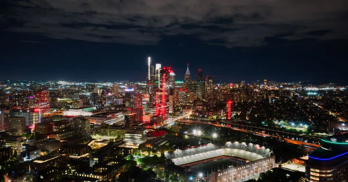 The Philadelphia skyline lit up at night, viewed across the city with a stadium in the distance.