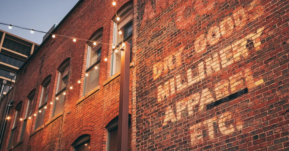 A restored brick warehouse at dusk, its wall painted with a faded vintage ghost sign and strung with cafe lights, the kind of old industrial building now filled with offices and studios.