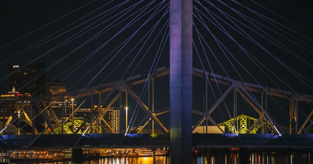 A cable-stayed bridge over the Ohio River in Louisville lit up at night, with city lights reflecting on the water.