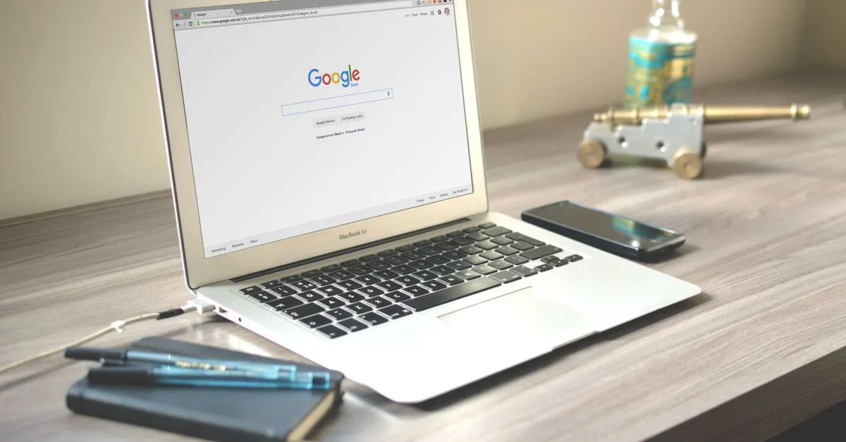 A laptop displaying a Google search on a wooden desk next to a smartphone and notebook.