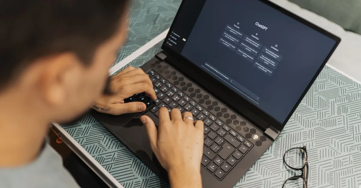 A man working on a laptop with an AI chat assistant open on the screen, wearing eyeglasses.
