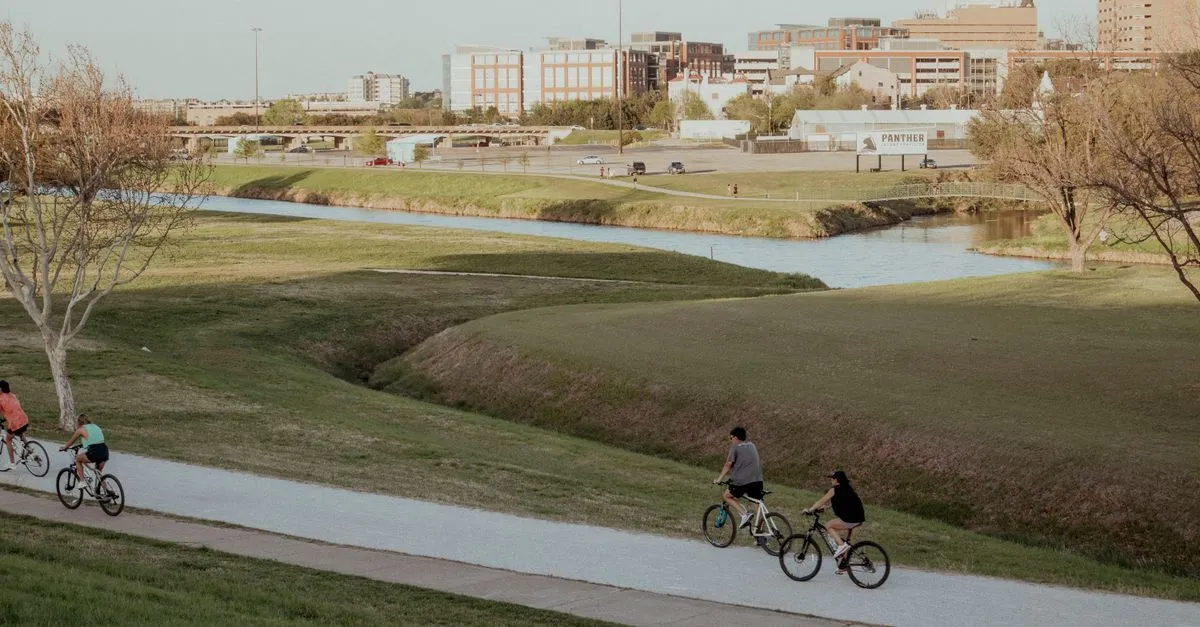 Cyclists on a paved riverside trail along the Trinity River, with Fort Worth's near-downtown skyline rising in the background.