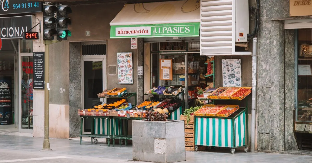 A small neighborhood grocery storefront with striped fruit and vegetable stands on the sidewalk.