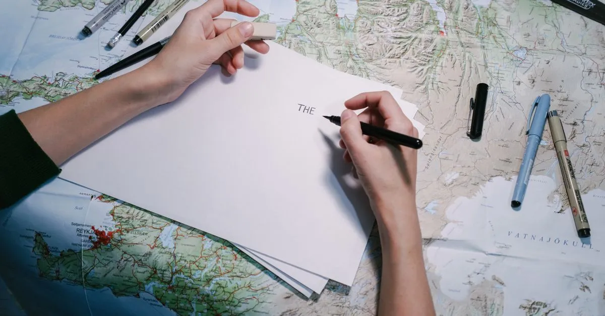 Hands writing on a sheet of blank paper resting on a detailed topographic map, surrounded by scattered felt-tip pens.
