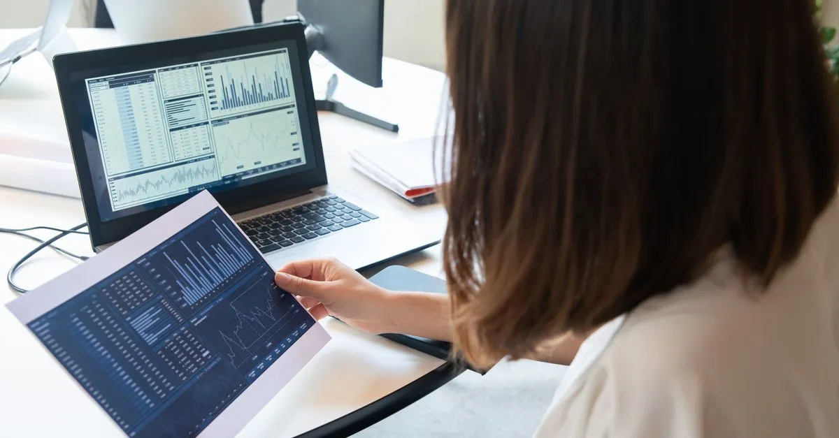 A person compares business charts on a printed sheet against the same data on a laptop screen, the way reputation monitoring compares what shows up across different sources.