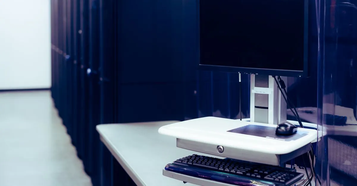 A row of dark server racks in a data center with a workstation monitor and keyboard at the end, the kind of hosting that answers requests quickly.