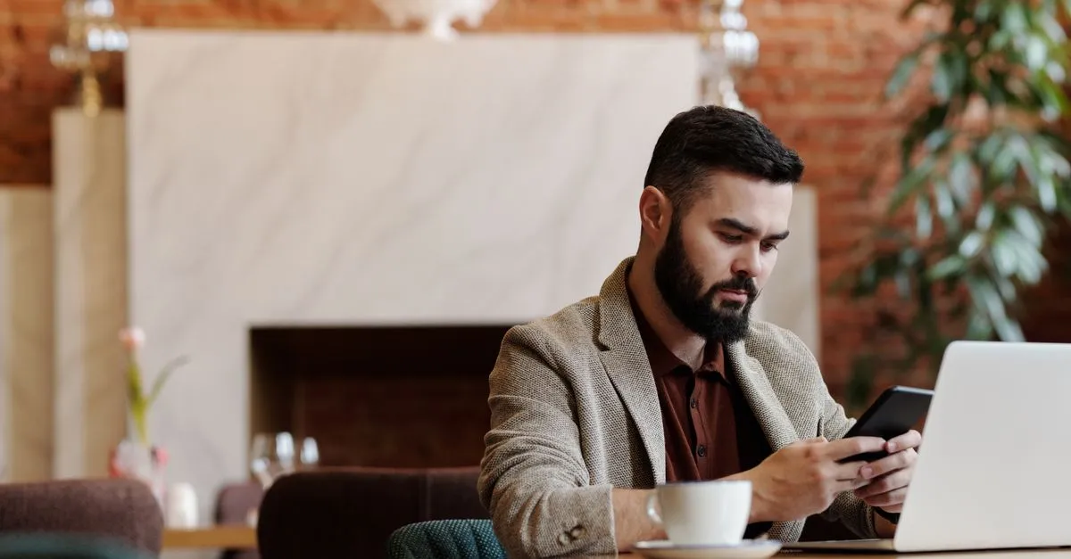 A man composing a reply on his phone next to a laptop in a coffee shop.