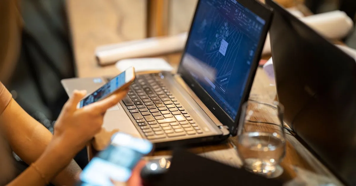 A person using a smartphone next to an open laptop on a wooden desk.