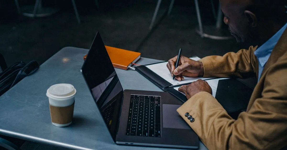 A man in a tan jacket writing notes beside an open laptop and a coffee cup at a metal table.
