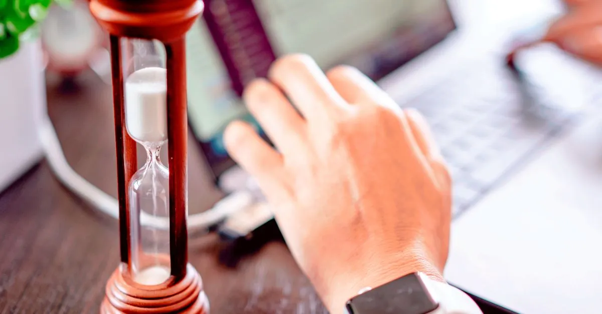 An hourglass with the sand running down sitting on a desk beside a hand reaching for a laptop keyboard, standing in for load time slipping away.