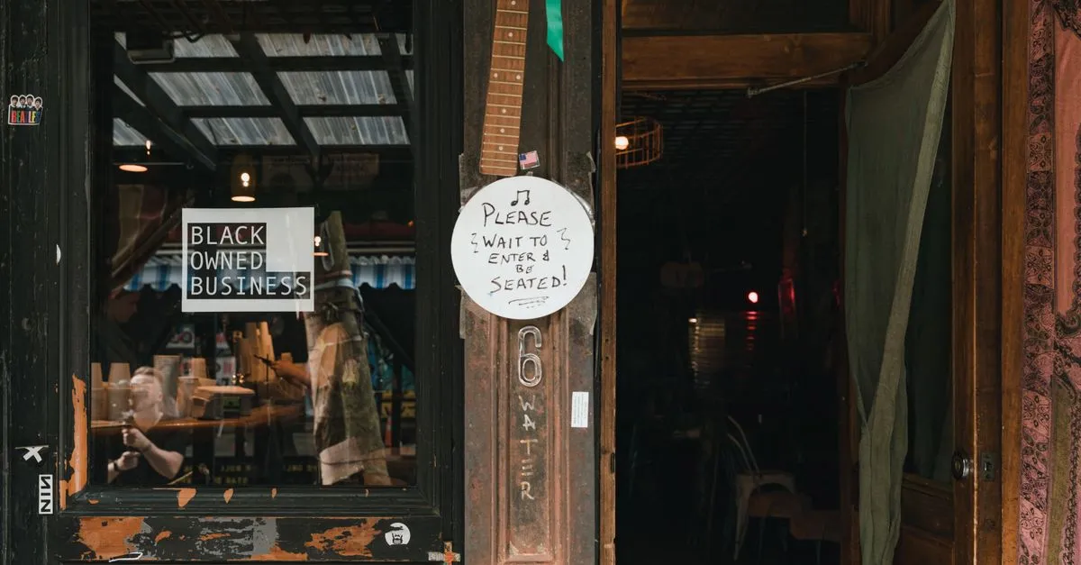 The weathered entrance of a cafe with a "Black Owned Business" sign in the window and a handwritten "Please wait to enter and be seated" sign beside the open door.