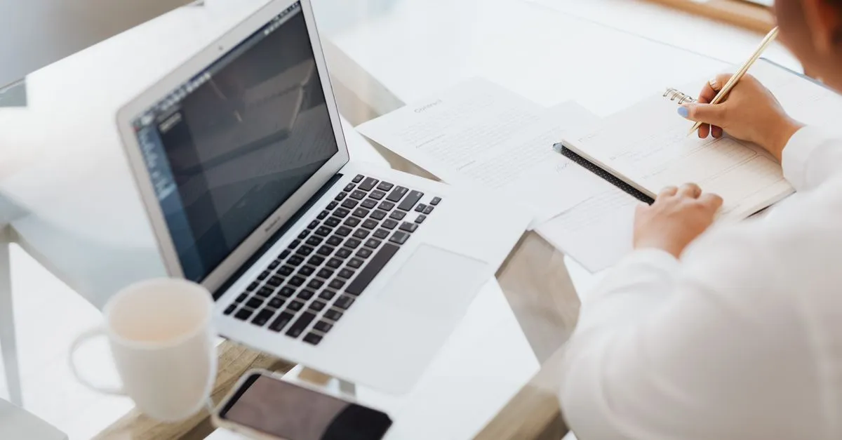 Person writing in a notebook next to a laptop and coffee mug, drafting page titles.